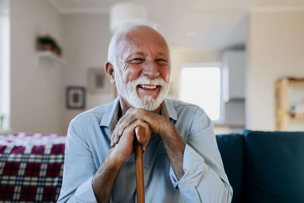 Portrait of happy senior man sitting at home with walking stock and smiling.Portrait of nice cheerful positive cheery stylish old man wearing checked shirt leaning on cane in white light modern interior studio room new house